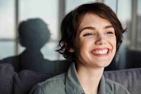 Image of brunette cute happy woman posing and laughing at camera while sitting on sofa at living roomの写真素材