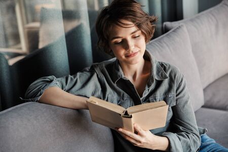 Image of brunette cute focused woman reading book while sitting on sofa at living room