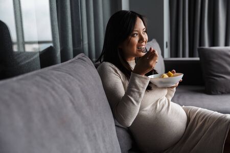 Photo of pleased pregnant asian woman eating on fruit salad while sitting on couch at homeの写真素材