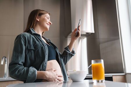 Photo of joyful pregnant woman with red hair using cellphone while having breakfast in modern kitchenの写真素材