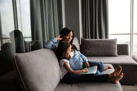 Photo of pleased pregnant asian woman with her little daughter reading book while sitting on couch at homeの写真素材