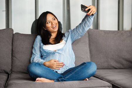 Photo of smiling pregnant asian woman taking selfie photo on mobile phone while sitting on couch at...