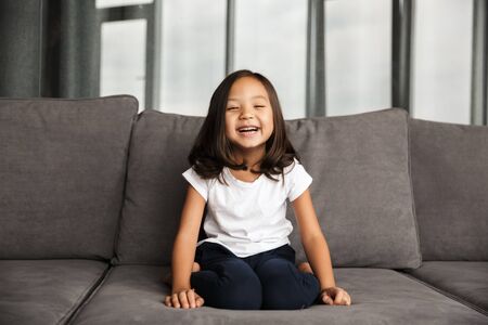 Photo of joyful little asian girl wearing casual wear laughing while sitting on couch at home