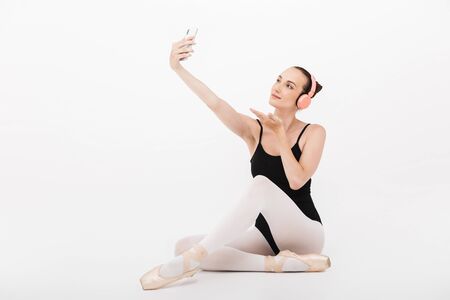 Image of caucasian young woman ballerina in headphones taking selfie photo on cellphone isolated over white wall backgroundの写真素材
