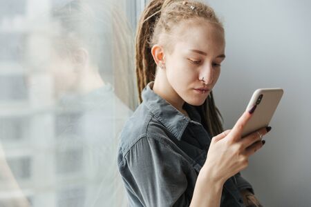 Image of a gorgeous concentrated young girl with dreadlocks and piercing indoors using mobile phone.