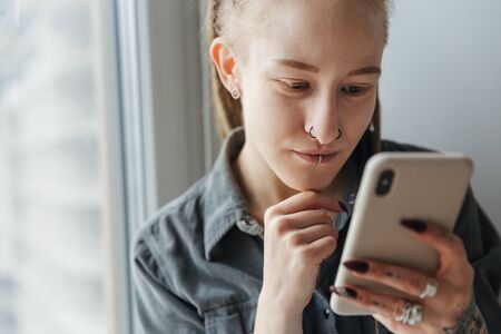 Image of a gorgeous concentrated young girl with dreadlocks and piercing indoors using mobile phone.の写真素材