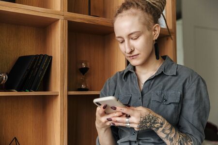 Image of a concentrated pretty young girl with dreadlocks and piercing indoors standing near shelf...