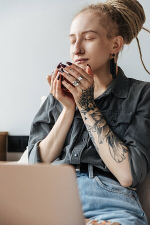 Image of a relaxing young girl with dreadlocks and piercing indoors using laptop computer drinking tea.の写真素材