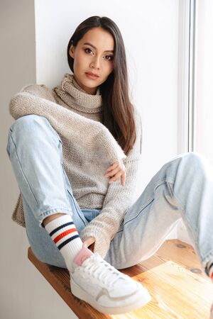 Image of beautiful asian woman looking at camera while sitting on window sill indoorsの写真素材
