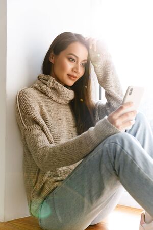 Image of beautiful asian woman typing on mobile phone while sitting on window sill indoorsの写真素材