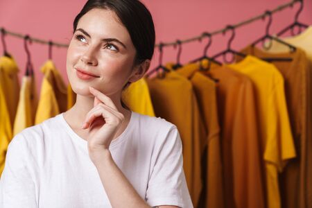 Photo of young thinking woman smiling and looking upward while standing at clothes rack in studioの写真素材