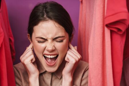 Photo of irritated cute woman plugging her ears and screaming while standing at clothes rack in...