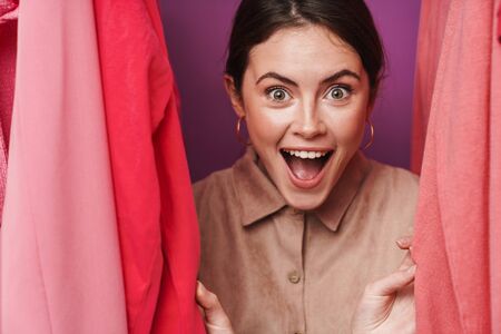 Photo of delighted cute woman smiling and looking at camera while standing at clothes rack in studioの写真素材