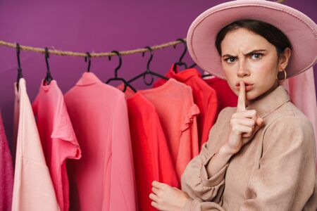 Photo of stylish displeased woman in hat making silence gesture while standing at clothes rack in studioの写真素材
