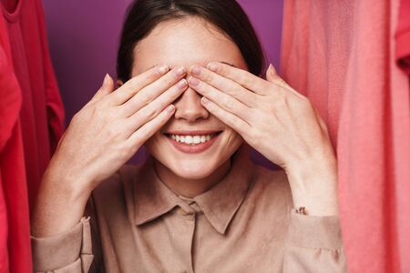 Photo of joyful beautiful cute woman covering her eyes while standing at clothes rack in studioの写真素材