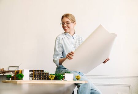 Photo of smiling blonde woman architect in eyeglasses working with drawings while designing draft at workplaceの写真素材