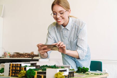 Photo smiling woman architect taking photo on cellphone while designing draft with house model at...