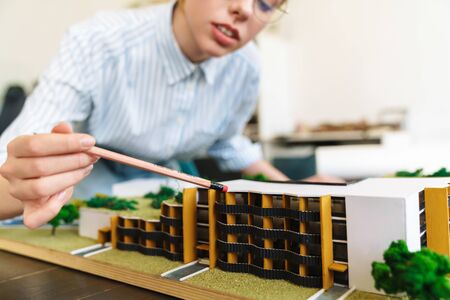 Photo of focused young woman architect in eyeglasses designing draft with house model and sitting at workplaceの写真素材