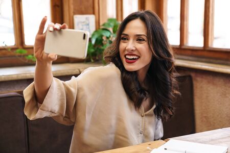Image of cheerful adult woman smiling while taking selfie on cellphone while sitting in cafe indoors