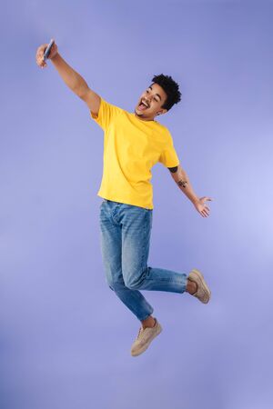 Photo of excited african american man jumping while taking selfie on cellphone isolated over purple backgroundの写真素材