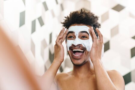 Photo of excited half-naked african american man with cosmetic mask on his face looking at mirror in...