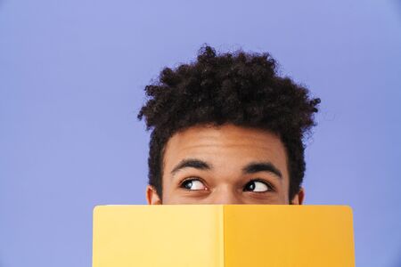 Photo of young african american man covering his face with exercise book isolated over purple...