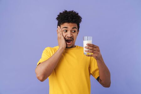 Photo of shocked african american man holding glass with milk isolated over purple backgroundの写真素材