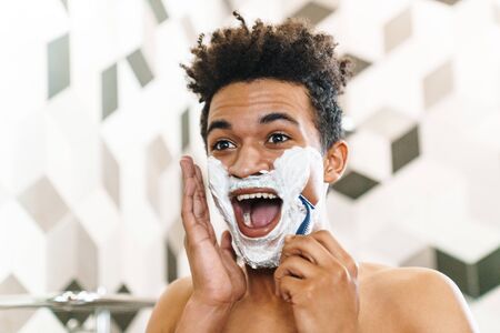 Photo of excited half-naked african american man shaving with foam while looking at mirror in bathroomの写真素材