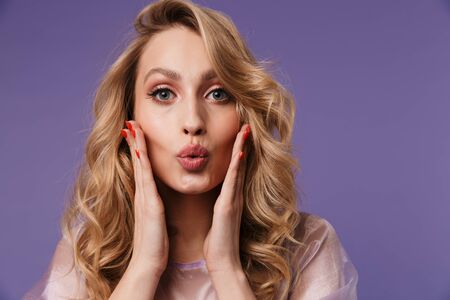 Image of young beautiful woman making kiss lips and touching her face isolated over purple backgroundの写真素材