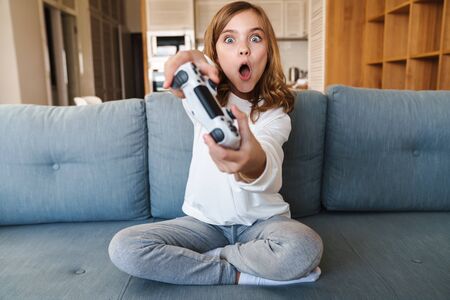 Photo of excited nice girl smiling and playing video game with joystick while sitting on couch at...