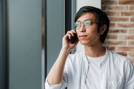 Image of handsome young asian man wearing eyeglasses talking on cellphone while standing by window in officeの写真素材