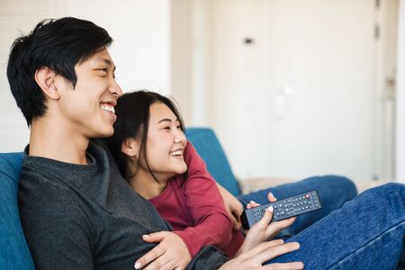 Photo of beautiful joyful asian couple watching tv and using remote control while sitting on sofa at...