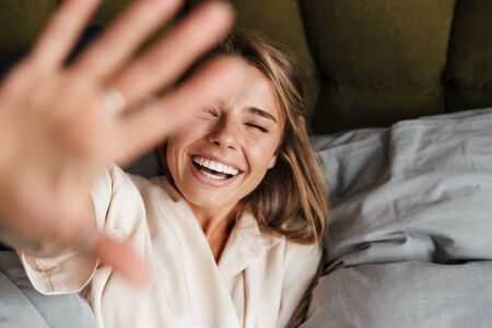 Image of laughing nice woman in pajama making fun and winking while lying in bed at homeの写真素材