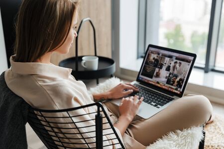 Image of focused blonde nice woman in stylish pajamas working with laptop while sitting on armchair at living roomの写真素材