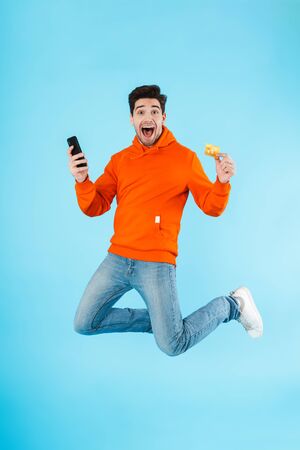 Portrait of a cheerful young man wearing hoodie jumping isolated over blue backgroundの写真素材