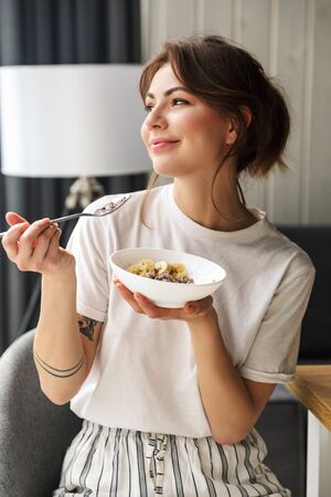 Photo of young smiling cute woman eating granola while having breakfast in cozy room at homeの写真素材
