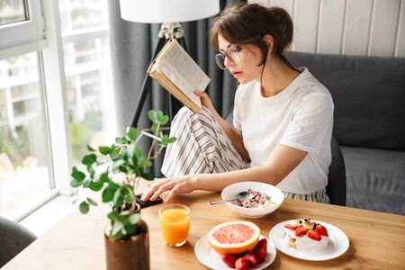 Photo of young woman reading book and using cellphone while having breakfast in cozy room at homeの写真素材