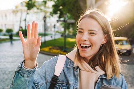 Beautiful cheerful young blonde woman wearing denim jacket walking in the city, waving handの写真素材