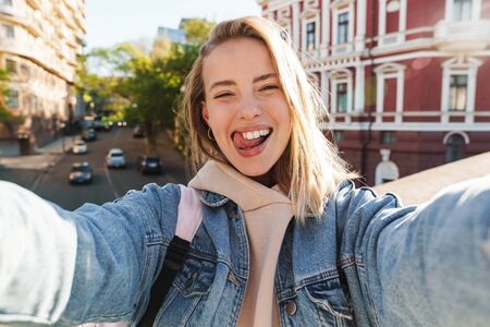 Beautiful cheerful young blonde woman wearing denim jacket walking in the city, taking a selfieの写真素材