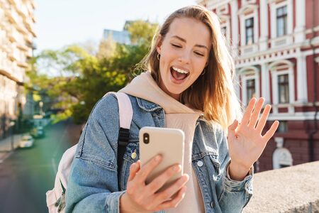 Beautiful cheerful young blonde woman wearing denim jacket walking in the city, taking a selfie, waving