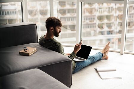 Image of focused young man using mobile phone and notebook while sitting on floor in living roomの写真素材