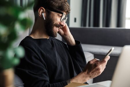 Image of laughing man in eyeglasses using cellphone and wireless earphones while sitting in living roomの写真素材