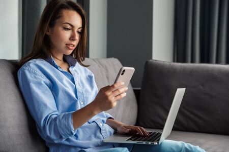 Image of young focused woman working with laptop and cellphone while sitting on couch at living room