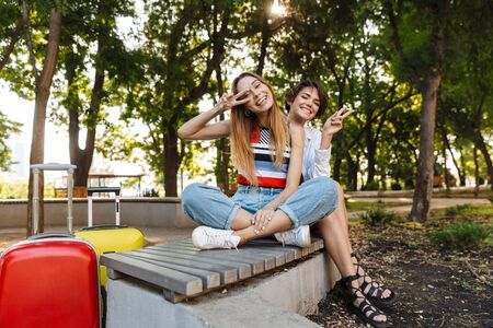 Photo of two delighted tourist women showing peace sign while sitting on bench with luggages in green parkの写真素材