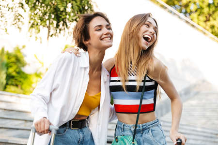 Photo of two excited tourist women rejoicing and laughing while walking with suitcases in parkの写真素材