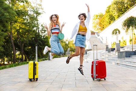 Photo of two attractive tourist women rejoicing and laughing while walking with suitcases in parkの写真素材