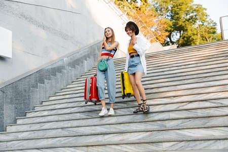 Photo of two happy beautiful women smiling while walking down stairs with suitcases at summer cityの写真素材