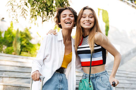 Photo of two beautiful tourist women rejoicing and laughing while walking with suitcases in parkの写真素材