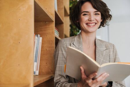 Smiling young woman entrepreneur reading book while standing in the office at the bookshelfの写真素材