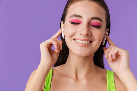 Photo of cute happy woman with bright makeup using earpods and smiling isolated over violet backgroundの写真素材